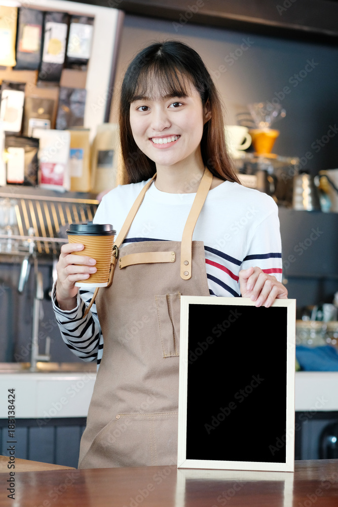 Young asia woman barista holding a diaposable coffee cup and blank menu chalkboard with smiling face at cafe counter background, small business owner, food and drink industry