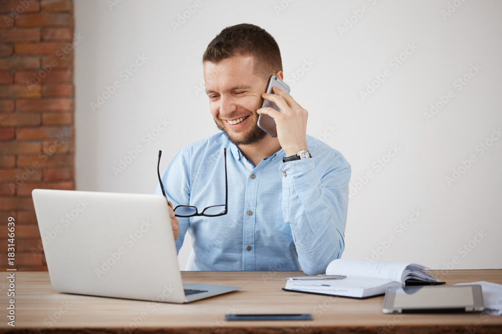 Cheerful professional good-looking shaved company director laughing, taking on glasses, looking in laptop monitor, talking on phone with manager.