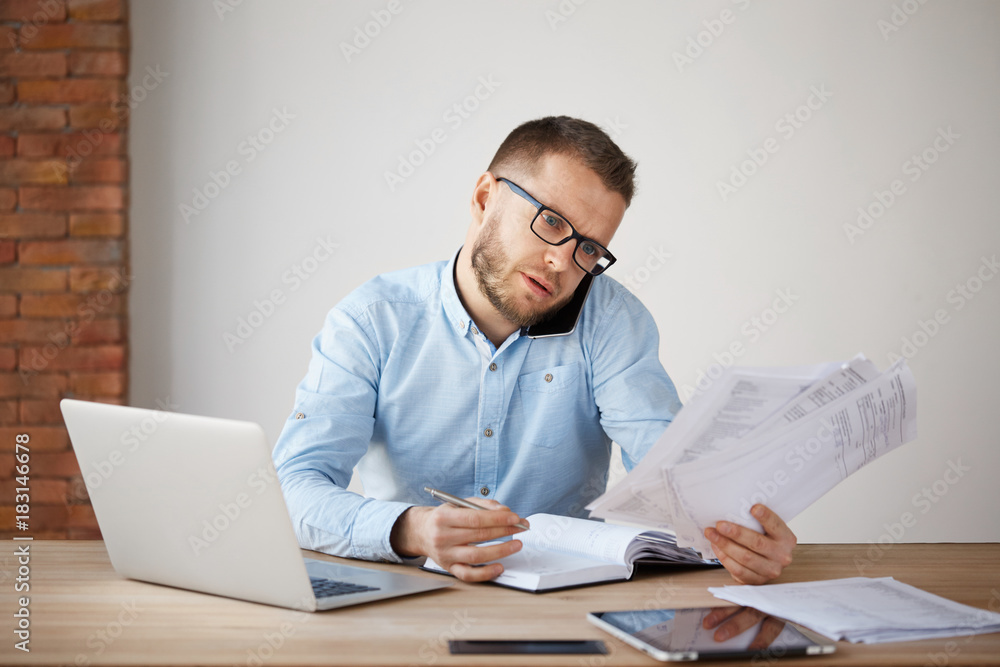 Busy concentrated unshaved businessman in glasses and shirt sitting in a comfortable light office, looking through papers, planning meeting with a team of workers by phone Busy concentrated unshaved businessman in glasses and shirt sitting in a comfortable light office, looking through papers, planning meeting with a team of workers by phone