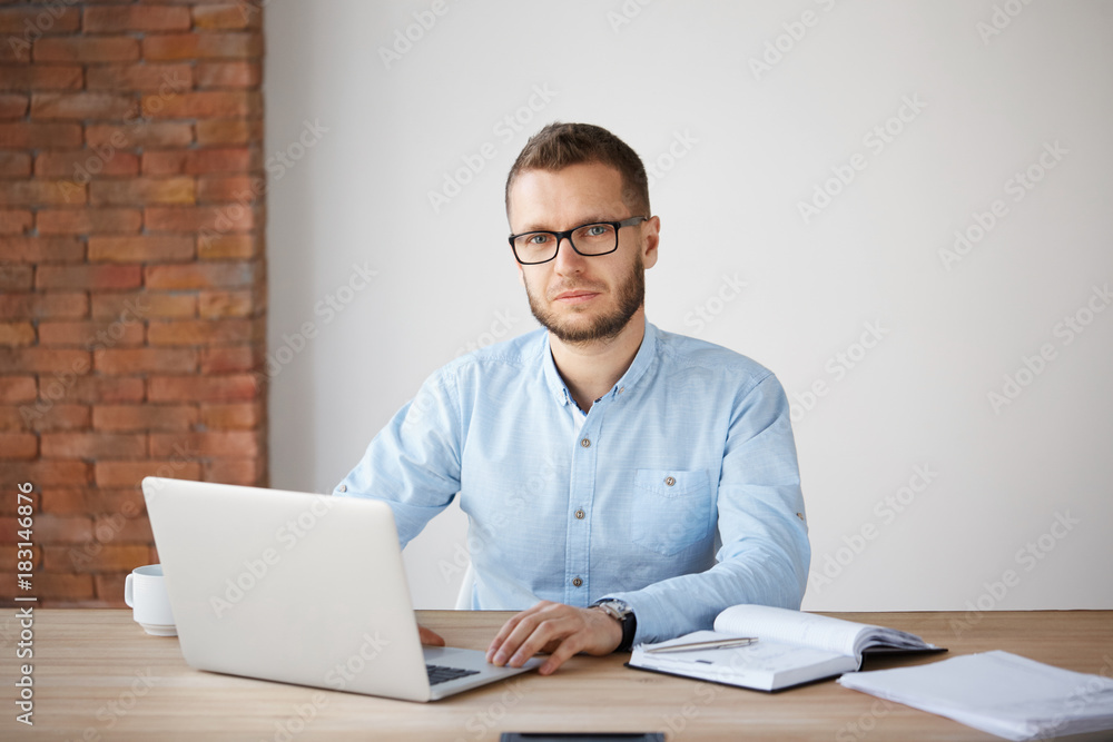 Portrait of serious mature unshaven businessman in glasses and blue shirt sitting at table, working on laptop computer, writing down tasks in notebook, looking in camera with relaxed expression. Portrait of serious mature unshaven businessman in glasses and blue shirt sitting at table, working on laptop computer, writing down tasks in notebook, looking in camera with relaxed expression.