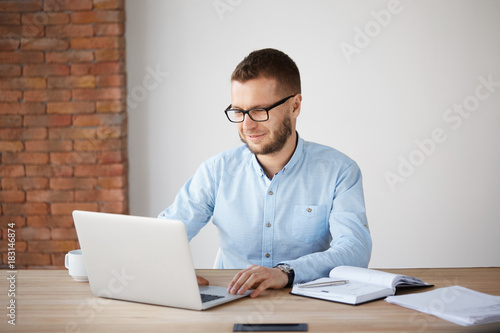 Portrait of cheerful handsome unshaven male company manager in glasses and casual clothes sitting at table in office, smiling gently, looking at laptop computer monitor, being happy to do favorite job