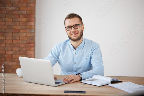 Close up of cheerful adult bearded male manager in glasses and classic shirt sitting at desk in office, working at personal computer, writing information in notebook, looking in camera with gentle