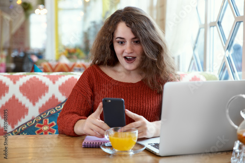 pretty young Caucasian red-haired girl student woman sitting at a table in a cafe with a Cup of sea buckthorn tea is holding a smartphone with a shocked surprised expression