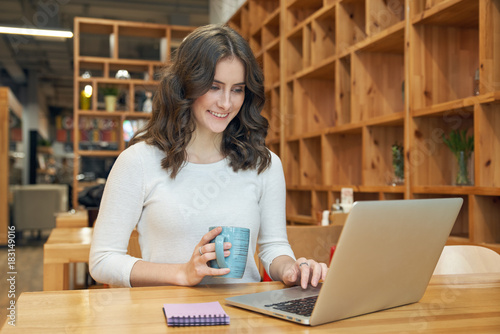 young attractive student girl doing homework sitting at a table with a mug of coffee and a laptop and talking on the social networks with her friends