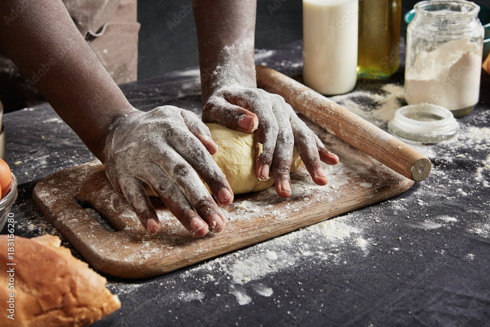 Indoor shot of black man with dirty hands kneads pasrty or dough carefully, uses rolling pin, prepares delicious pizza for guests or relatives, wants to be praised for his culinary abilities. Cooking