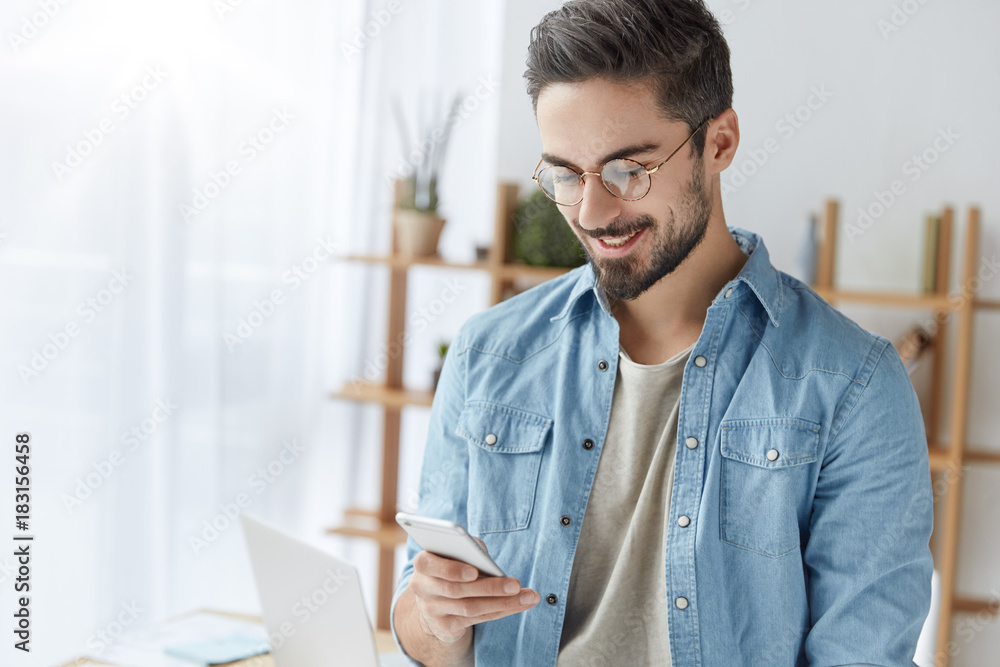 Fototapeta premium Cheerful young bearded male starts own business, recieves messages of encouragement from friends, who support and believe in his success, uses modern smart phone, texts and chatts with people