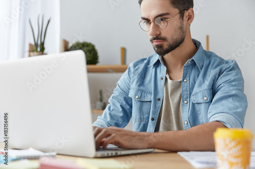Serious male model wears denim shirt and glasses, checks emails online, uses modern laptop computer, works remotely on project, uses free wifi, keeps hands on keyboard, communicates with partners