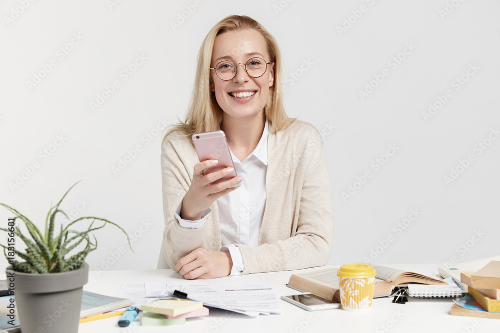 Portrait of cheerful blonde young European female sits at workplace, creats new innovative project, holds smart phone, messages with friends, agrees about meeting, uses free internet connection Portrait of cheerful blonde young European female sits at workplace, creats new innovative project, holds smart phone, messages with friends, agrees about meeting, uses free internet connection