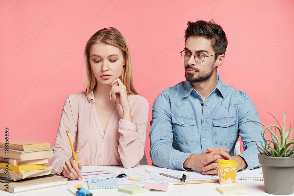 Indoor shot of creative female and male workers sit next to each other, surrounded with papers and books, focused on work, isolated over pink background. People, work, education, cooperation concept Indoor shot of creative female and male workers sit next to each other, surrounded with papers and books, focused on work, isolated over pink background. People, work, education, cooperation concept