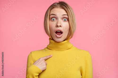 Shocked stunned light haired woman points or indicates at herself with fore finger, surprised be accused of fault or thing she didn`t do, proves rightness. Frightened terrified female model. photo