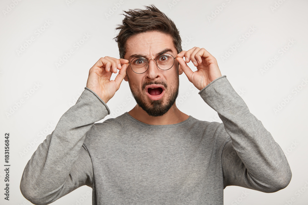 Fototapeta premium Puzzled bearded man wonders about something, finds out results of test or his fault, looks through spectacles, has trendy hairdo, wears casual grey sweater, isolated over white studio background