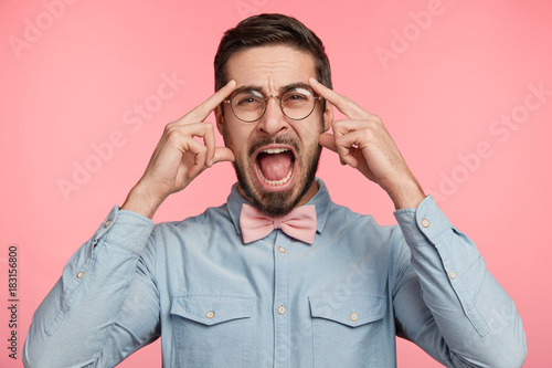 Young bearded Caucasian male screams loudly, keeps fingers on temples as has bad headache after working all night, expresses negative emotions and feelings, poses alone over pink background.
