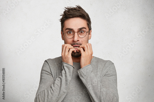 Indoor shot of nervous worried handsome man bites fingers, being scared of horror film or afraid of exams, has frightened expression, isolated over white concrete background. Emotional adult photo