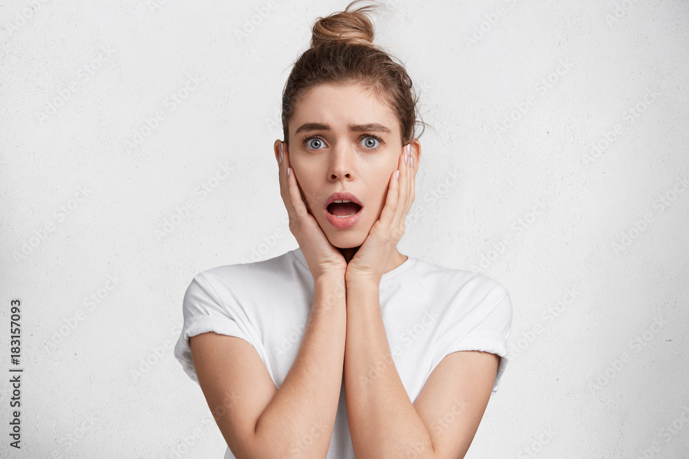 Surprised young pretty female has hair tied in knot, looks with opened mouth, shocked to see something awful, dressed casualy, isolated over white background. People, surprisment, facial expresions Surprised young pretty female has hair tied in knot, looks with opened mouth, shocked to see something awful, dressed casualy, isolated over white background. People, surprisment, facial expresions
