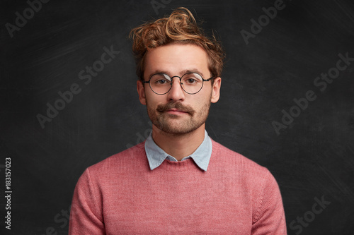 Horizontal shot of confident pleasant looking intelligent male nerd wears elegant clothes, round optical spectacles, poses against chalk blackboard, prepares for presenting information to audience