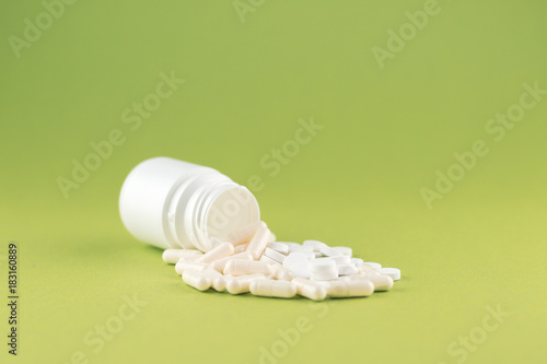 Close up white pill bottle with spilled out pills and capsules on olive yellow background with copy space. Focus on foreground, soft bokeh. Pharmacy drugstore concept
