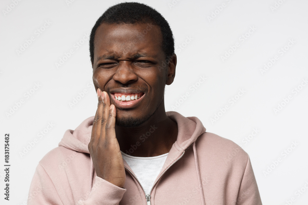 Indoor portrait of young worried African American male isolated on white background dressed in white T-shirt and pink hoodie experiencing strong toothache pressing hand to chin because of sore jaws