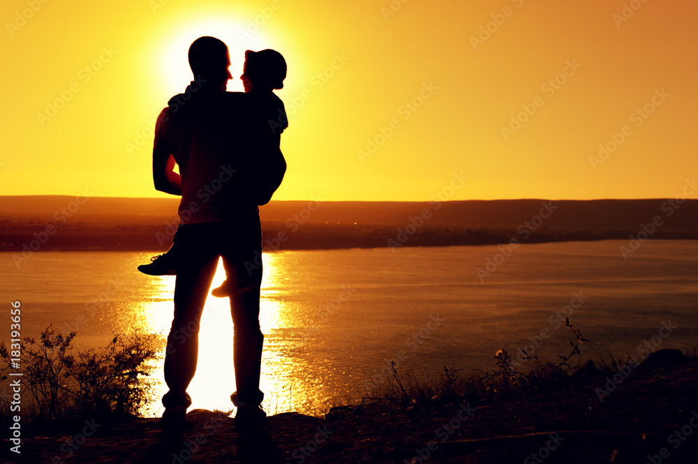 Rear view -  silhouette of the father holding his little son in arms and admiring the sea view on sunny summer evening