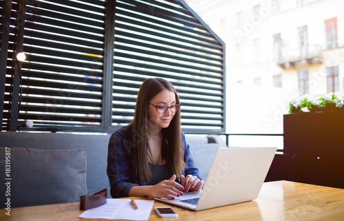 Young smart woman in glasses developer wed sites working on laptop computer in restaurant. Beautiful female successful freelancer using net-book for work. Pretty hipster girl keyboarding on notebook