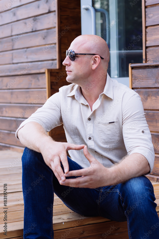 Brutal Bald man sit sits on the steps on the wooden background, nature shirt, jeans, sunglasses, after barbershop, enjoying sun at start spring, outdoor, summer