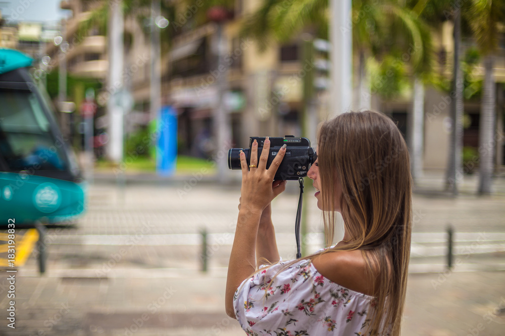 Tenerife. Spain. Cute blonde girl is observing and  filming streets in Santa Cruz de Tenerife with colourful tram on the background. Girl travels concept. Travel to Canary Islands.