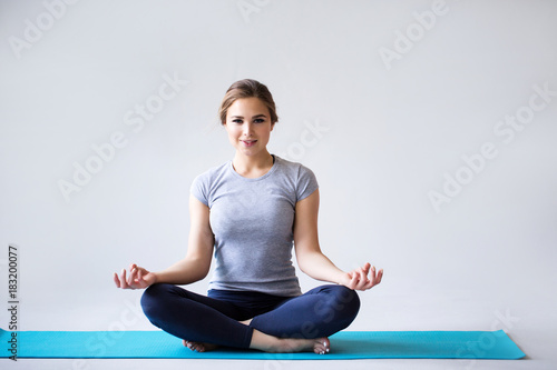 Beautiful young woman in sportswear practicing yoga while sitting in lotus position on gray background.