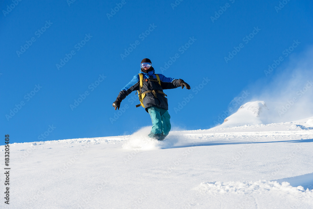 Freeride snowboarder rolls on a snow-covered slope leaving behind a snow powder against the blue sky