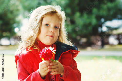 Cute girl blonde in a red jacket holding a flower in the Park outdoors