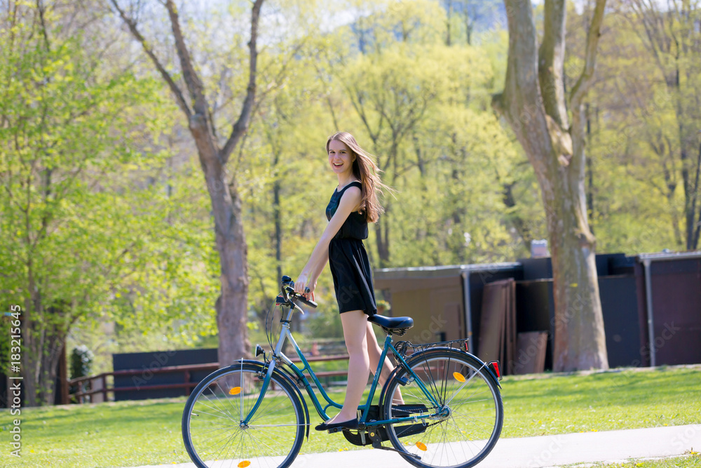 Portrait view of girl on bicycle wearing on black short dress. Young happy Woman riding along road on green spring outdoor Park. Sporty young girl riding a bicycle on a sunny morning Portrait view of girl on bicycle wearing on black short dress. Young happy Woman riding along road on green spring outdoor Park. Sporty young girl riding a bicycle on a sunny morning