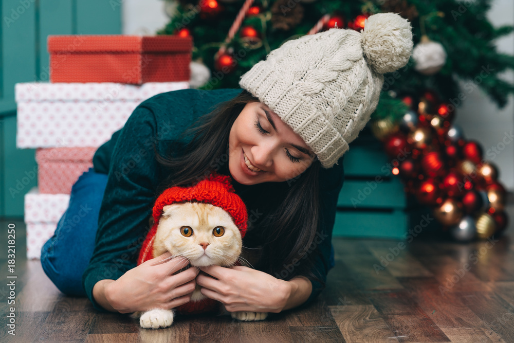 Portrait of young woman and cute cat wearing sweater and knitted hats. christmas tree and presents background Portrait of young woman and cute cat wearing sweater and knitted hats. christmas tree and presents background