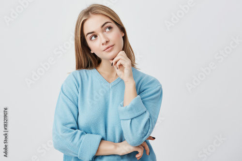 Face expressions and emotions. Thoughtful young pretty girl in blue sweater holding hand under her head, having doubtful look while can't decide what clothes to wear on friend's birthday party
