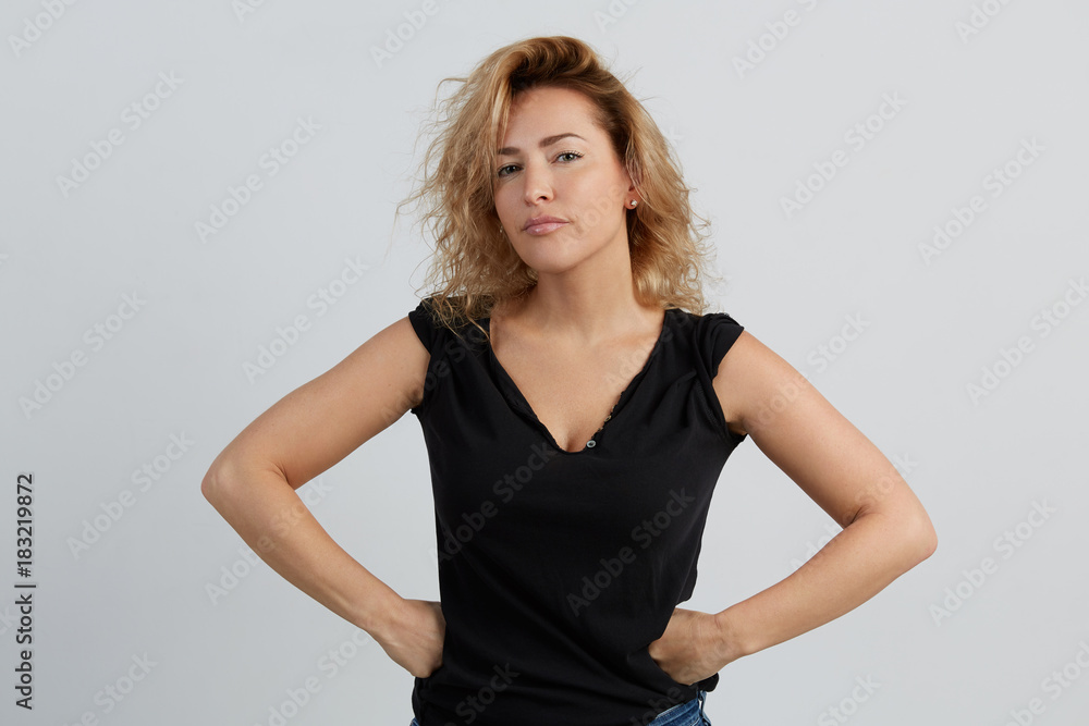 Emotional beautiful young adult woman portrait. Studio shoot. Isolated on White Background. A woman in a black T-shirt with a deep neckline looks severely. Her hands are pinned on her hips.