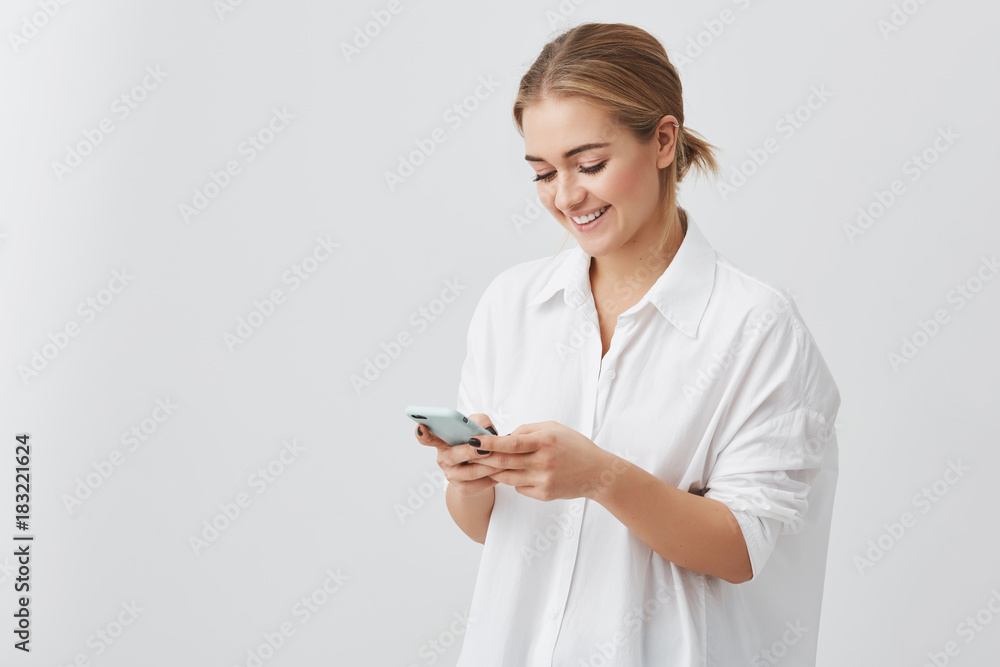 Attractive pretty woman with fair hair in white shirt smiling while using cell phone chatting with her boyfriend posing against gray studio wall. Beauty and youth concept Attractive pretty woman with fair hair in white shirt smiling while using cell phone chatting with her boyfriend posing against gray studio wall. Beauty and youth concept