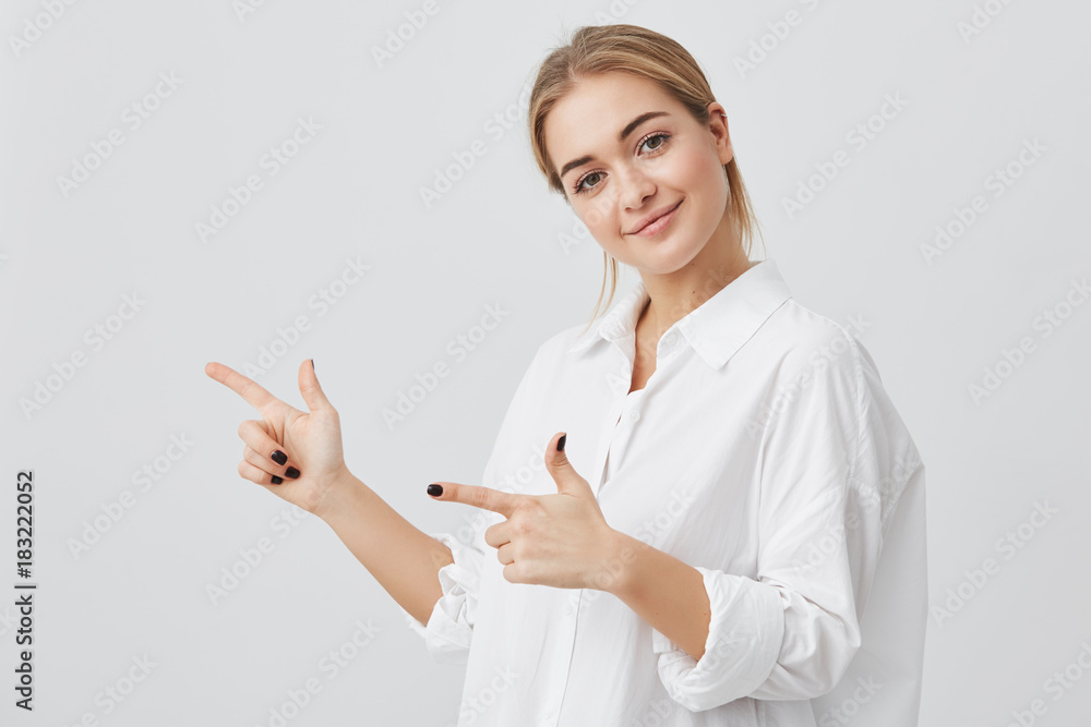 Woman smiling cheerfully and pointing her index fingers, showing copy space for your text or advertising content. Studio shot of attractive young blonde girl isolated on gray background Woman smiling cheerfully and pointing her index fingers, showing copy space for your text or advertising content. Studio shot of attractive young blonde girl isolated on gray background