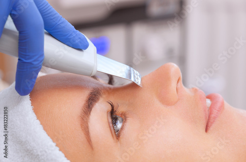 The doctor-cosmetologist makes the apparatus a procedure of ultrasound cleaning of the facial skin of a beautiful, young woman in a beauty salon. Cosmetology and professional skin care.
