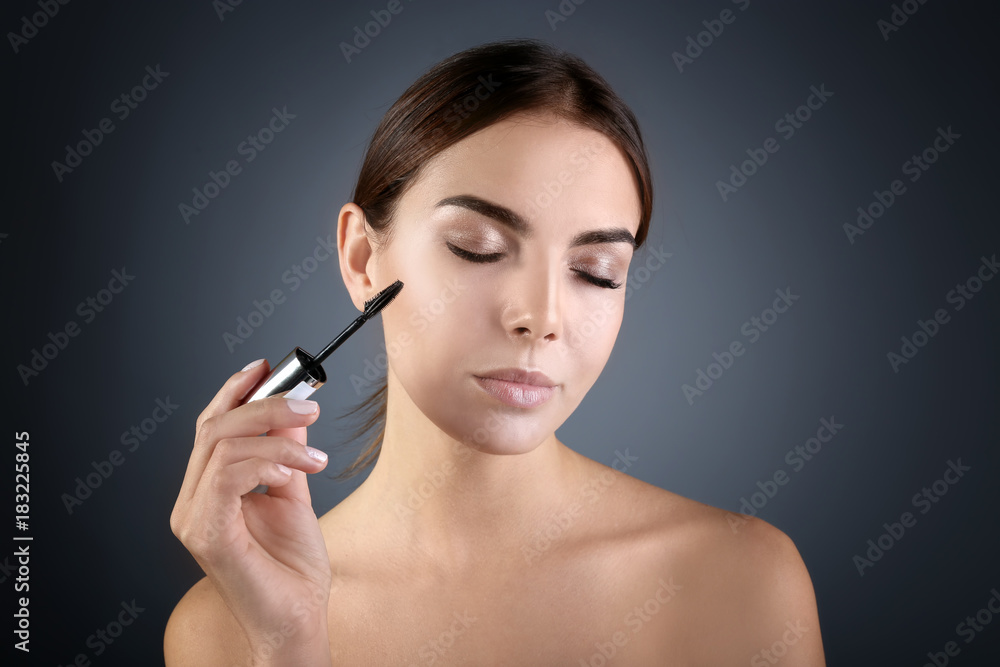 Obraz premium Beautiful young woman with eyelash extensions applying mascara against grey background