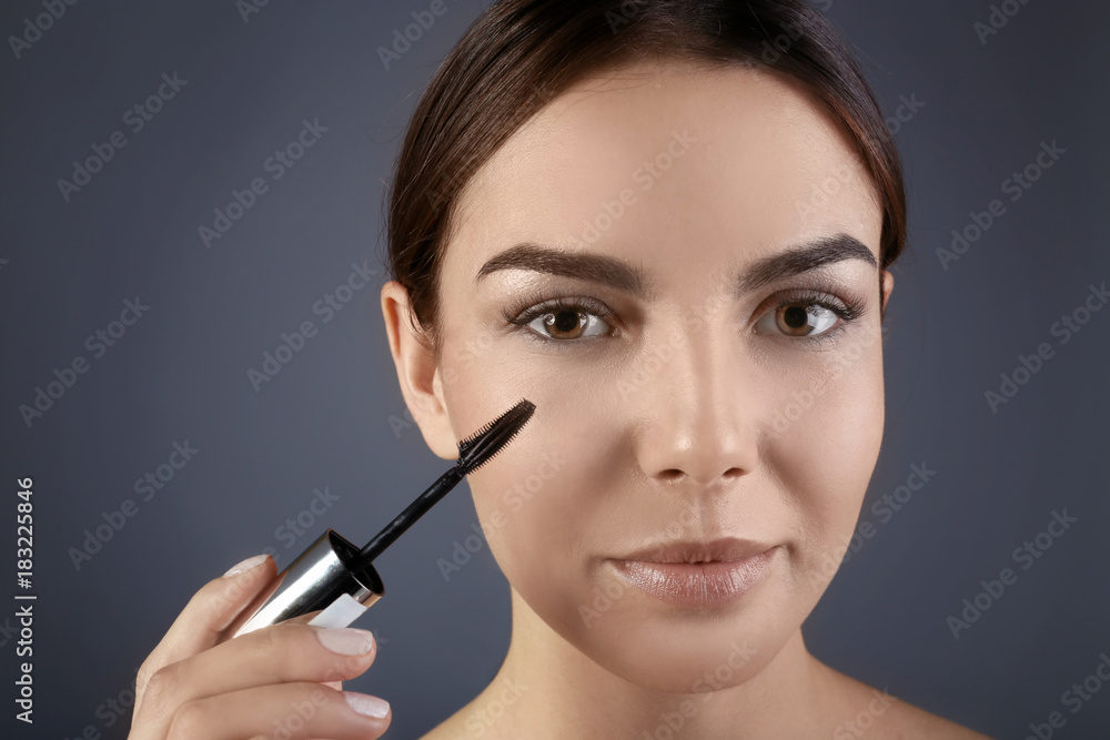 Beautiful young woman with eyelash extensions applying mascara against grey background Beautiful young woman with eyelash extensions applying mascara against grey background