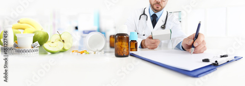 Nutritionist doctor writes the medical prescription for a correct diet on a desk with fruits, drugs and supplements, web banner and copy space template photo