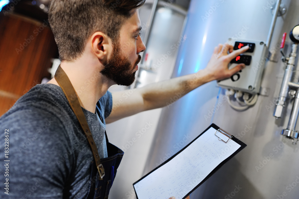 Fototapeta premium A handsome bearded male brewer records data against a background of beer tanks. Brewing