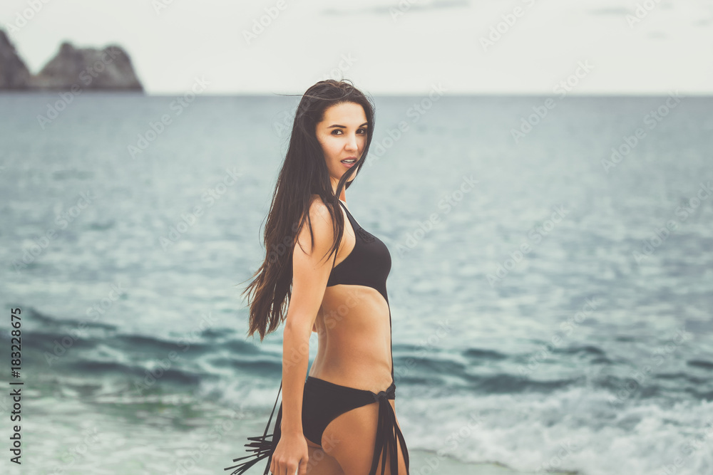 Young beautiful playful woman on the beach, ocean and mountain background. She is dressed in black swimwear. hipster style, soft light Young beautiful playful woman on the beach, ocean and mountain background. She is dressed in black swimwear. hipster style, soft light