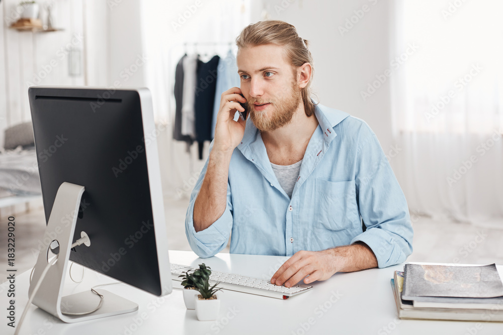 Bearded skilled young fair-haired businessman works on new project, sits in front of screen, has phone conversation, discusses financial report with businesspartner. Office worker chats with boss