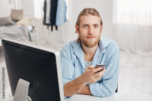 Bearded good-looking male office worker with gentle smile reads notification on smart phone, sits in front of screen at coworking space with cell phone, sends feedback to coworkers, browses internet