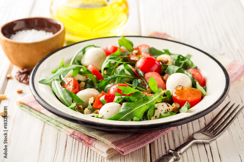 Fresh salad with arugula, cherry tomatoes, mozzarella cheese and walnuts on white wooden background.