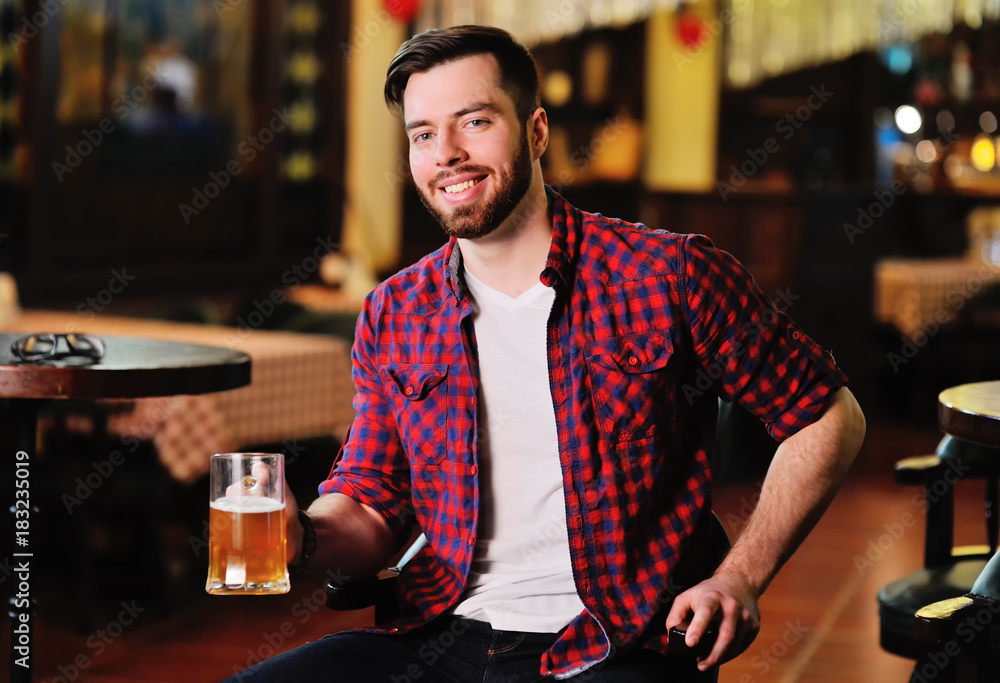 Cute young man in a plaid shirt drinking beer sitting on a chair in a pub
