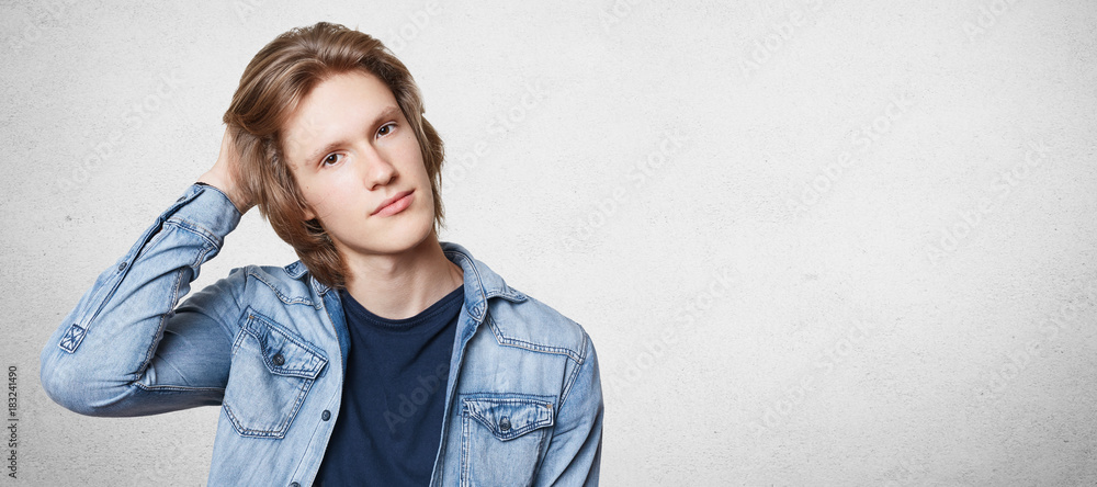 Horizontal portrait of fashionable young guy with appealing appearance, dressed in stylish clothes, poses for magazine cover, stands against copy blank concrete wall with copy space for advertisment