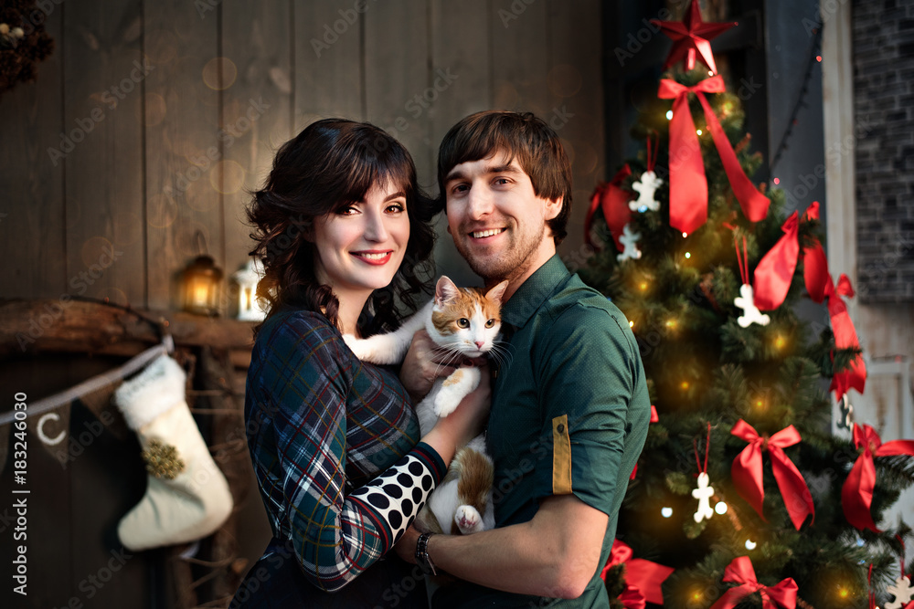 Beautiful couple in love in green with a red cat on a background of Christmas tree decorations. Happy family in new year loft with red yellow bokeh. Wooden wall. Copy space. Close up portrait. Beautiful couple in love in green with a red cat on a background of Christmas tree decorations. Happy family in new year loft with red yellow bokeh. Wooden wall. Copy space. Close up portrait.
