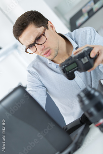 Spectacled man holding camera looking at laptop