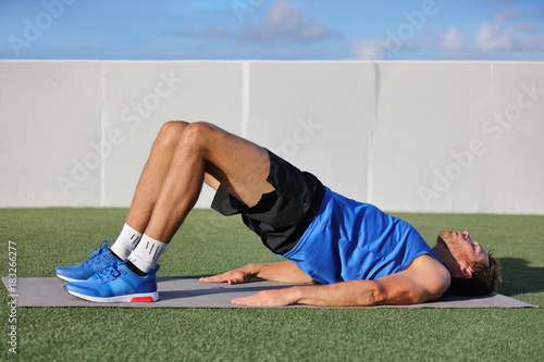 Fitness man doing bodyweight glute floor bridge pose yoga exercise. Fit athlete exercising glutes muscles with butt raise at summer outdoor gym instruction class on grass.