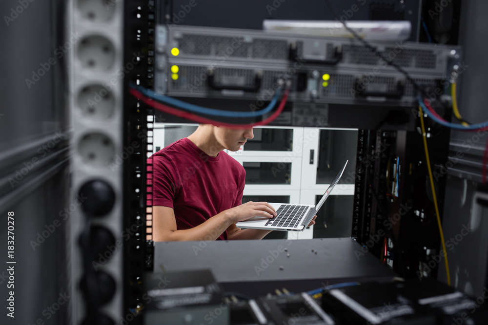 Portrait of modern young man holding laptop standing in server room working with supercomputer Portrait of modern young man holding laptop standing in server room working with supercomputer