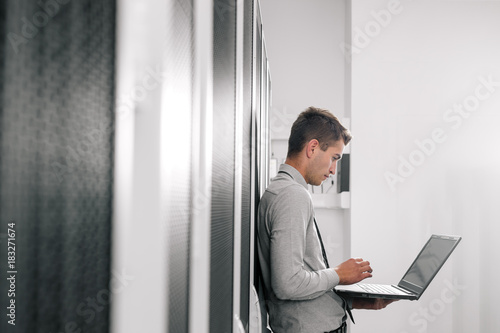 Portrait of modern young man holding laptop standing in server room working with supercomputer